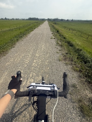A cyclist’s view over black drop handlebars along a straight gravel road between green fields on the way to Drumheller.