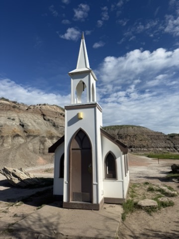 Drumheller’s tiny white Little Church, with its pointed steeple framed by layered badlands hills and blue sky.