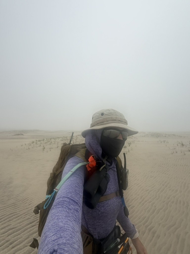 Weronika in field gear hiking across rippled sand dunes in dense fog.