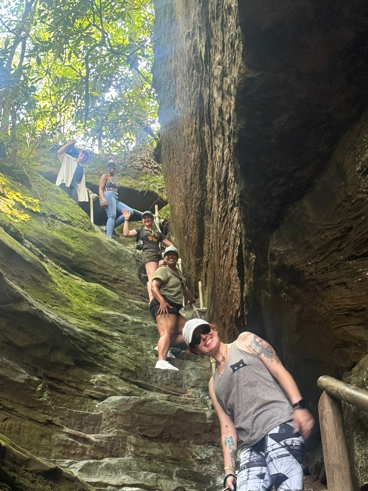 A group of workshop colleagues smiling and waving on a narrow stone stairway between rock walls on the Original Natural Bridge Trail.